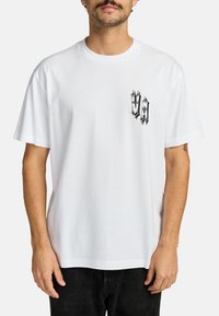 Witte katoenen t-shirt met een zwart grafisch ontwerp op de linkerkant van de borst. Loose fit met korte mouwen en een ronde hals.