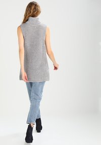 Grå stickad ärmlös tunika med hög krage, ihop med ljusblå jeans och svarta ankelstövlar, visad bakifrån.