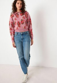 Roze gebreide trui met bloemenpatroon, gecombineerd met lichtblauwe jeans en witte sneakers. De trui heeft een geribbelde zoom en manchetten.