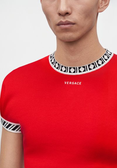 Άνδρας που φοράει κόκκινο T-shirt Versace με μαύρη και λευκή διακοσμητική λεπτομέρεια στη λαιμόκοψη και στις άκρες των μανικιών.