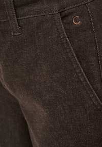 Gros plan sur un jean en denim marron, montrant la ceinture, la poche avant, le passant de ceinture et un petit logo métallique "C" au-dessus de la poche.