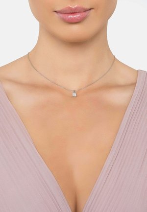 Collana di catena argento con un piccolo ciondolo quadrato che presenta una pietra preziosa trasparente. Indossata sopra una maglietta rosa chiaro con texture a pieghe.