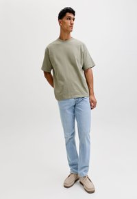 Tânăr stând cu mâinile la spate, purtând un tricou verde deschis oversized, blugi albaștri deschiși și pantofi bej din piele întoarsă, pe un fundal alb.