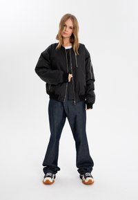 Black bomber jacket with zip front, ribbed cuffs, and side pockets, paired with loose-fitting dark denim pants and multi-colored sneakers.