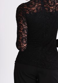 Sort blonde top med lange ærmer, indviklede blomster mønstre og en tætsiddende silhuet; struktureret stof med en gennemsigtig ryg.