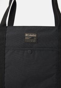 Sac tote noir en tissu durable, de forme rectangulaire, avec des poignées supérieures et une étiquette de marque en lettres dorées et un bouton-pression.