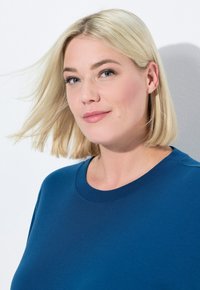 T-shirt di cotone blu con scollatura tonda, maniche corte e texture liscia. Il modello ha capelli biondi lisci ed è posato su uno sfondo chiaro.