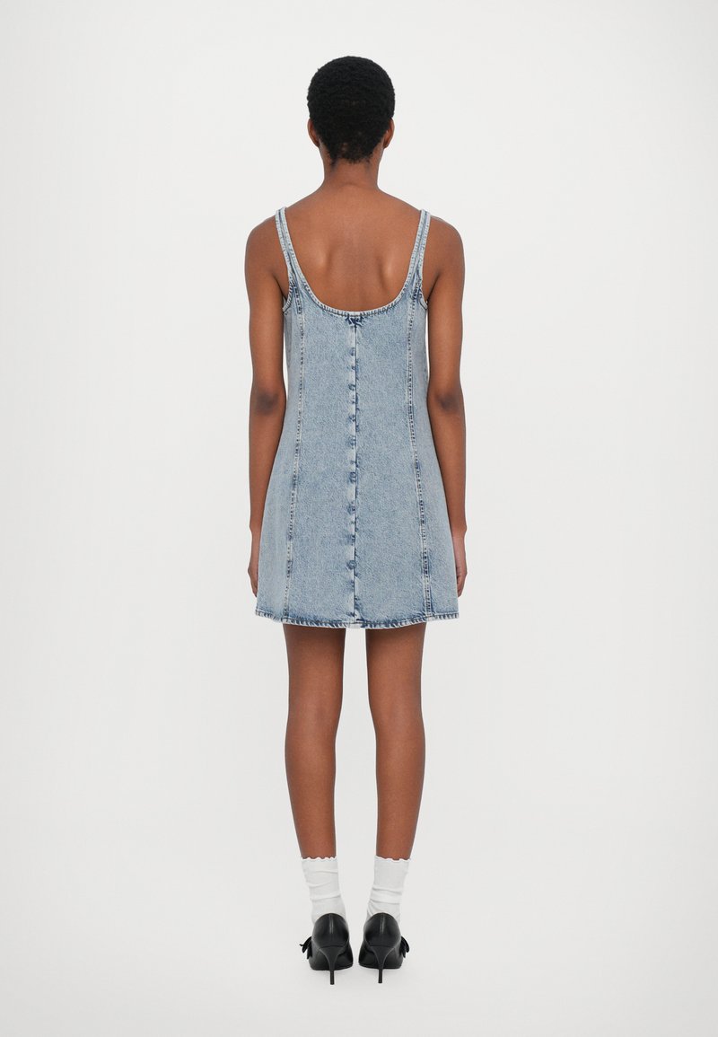 Jeanskleid, hellblau, ärmellos mit rundem Rückenausschnitt, hat eine durchgehende Knopfleiste und einen ausgefransten Saum. Getragen mit weißen Socken und schwarzen High Heels.