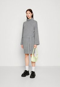 Robe grise longueur genou avec un col montant, des manches longues et une taille ceinturée. Associée à un sac à main vert clair et des chaussures à plateforme noires épaisses.