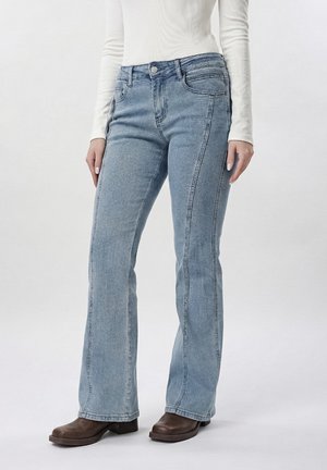 Femeie purtând blugi bleu deschis cu croială bootcut, bluză albă cu mânecă lungă și cizme maro până la gleznă, stând în picioare pe un fundal simplu.