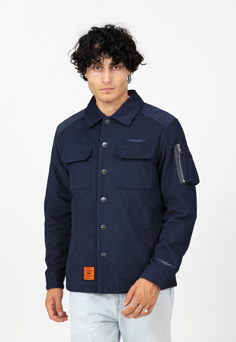 Marineblauwe jas van een getextureerde stof, met meerdere zakken, knoopsluiting aan de voorkant en een oranje label aan de zoom; heeft een ritszak op de mouw.