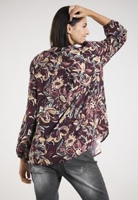 Langärmlige Blumenshirt in lila, beige und grünen Mustern. Hergestellt aus leichtem Stoff mit lockerer Passform und elastischen Bündchen.