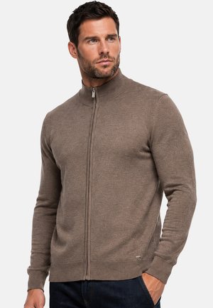 Uomo con capelli corti e scuri che indossa un maglione marrone con zip e pantaloni blu scuro, in piedi con una mano in tasca, guarda verso destra.