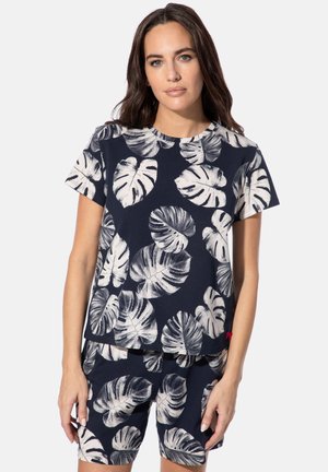 Frau trägt passendes dunkelblaues Hemd und Shorts mit großen weißen tropischen Blattmustern, steht vor einem einfarbigen hellen Hintergrund.