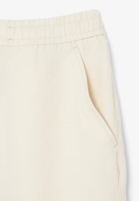 Close-up van beige stoffen broek met een elastische tailleband en een zijzak met zichtbare stikdetails.
