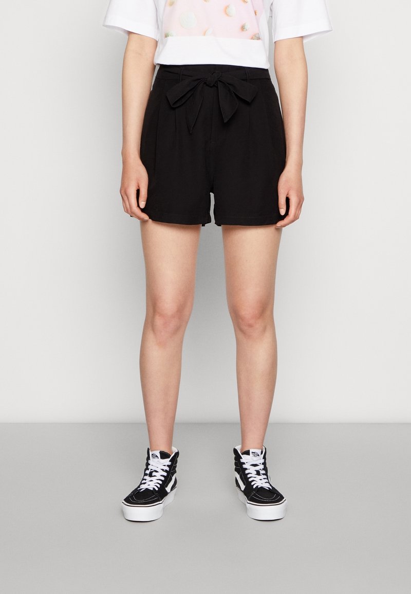 Schwarze Shorts aus leichtem Stoff mit einem gebundenen Gürtel in der Taille. Kombiniert mit schwarz-weißen High-Top-Sneakern.