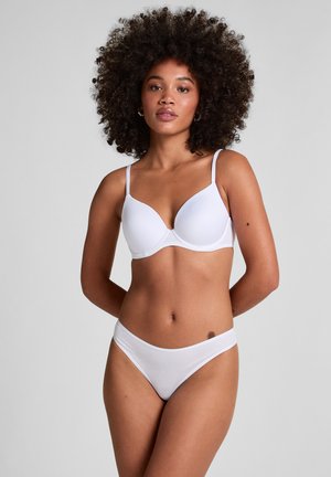 Donna con capelli ricci che indossa un reggiseno bianco e biancheria intima coordinata, in piedi con le mani dietro la schiena davanti a uno sfondo chiaro e semplice.