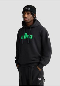 Felpa nera con logo "elko" verde, cappuccio con cordino, polsini a coste, abbinata a pantaloni neri con tasche con zip laterali e patch del logo.
