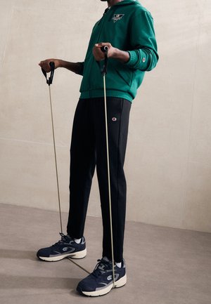 Person in grünem Kapuzenpullover und schwarzer Hose, die ein Widerstandsband unter den Füßen für eine Oberkörperübung an einer beigen Wand benutzt.