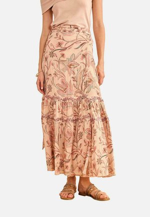 Mujer con una falda maxi rosa con estampado floral y volantes en capas, sandalias marrones con tachuelas y una blusa sin mangas beige sobre un fondo liso.