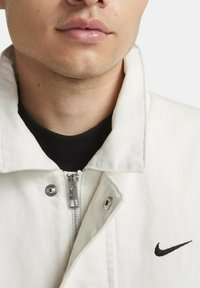 Chaqueta de mezclilla blanca con cierre de cremallera, herrajes plateados y un logo negro de Nike en el lado izquierdo del pecho. Tejido texturizado con un cuello clásico.