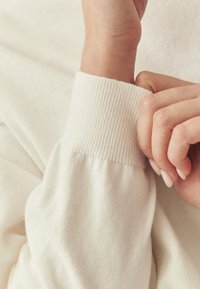 Pull en maille de couleur crème avec un poignet côtelé. Le tissu semble doux, avec une texture lisse et un léger éclat.