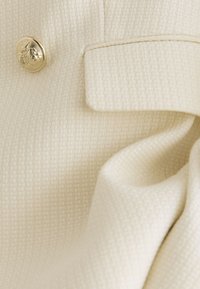 Krémová textúrovaná látka so zlatým dekoratívnym gombíkom a obdĺžnikovým látkovým prekrytím, naznačujúca záber detailu na mieru šitého odevu.