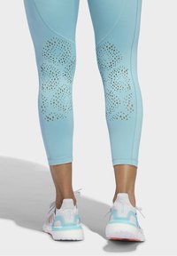 Leggings sportivi azzurri chiari con motivo perforato dorato nella parte inferiore delle gambe, abbinati a sneakers bianche con accenti blu pastello.