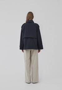Navy blue coat with broad lapels, back flap detail, and long sleeves, paired with light beige wide-leg trousers. Smooth texture, minimal design.