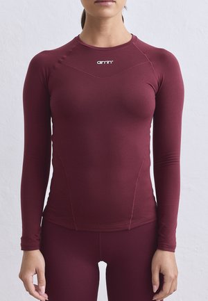 Femme portant un haut de sport bordeaux à manches longues ajusté et un legging assorti sur un fond clair uni.