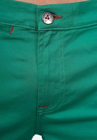 Pantaloni verdi realizzati in tessuto morbido, con chiusura a pulsante tondo, dettagli di cucitura rossa e un primo piano della zona della vita.
