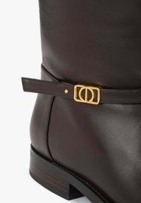 Brązowy skórzany but z zaokrąglonym noskiem, ozdobiony złotym klamrą i paskiem. Obcas jest niski i kwadratowy, z delikatną fakturą.