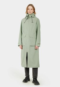 Gabán de lluvia largo en verde claro con capucha, cierre frontal y dos grandes bolsillos de parche. Textura suave, diseño minimalista, con cordones ajustables.