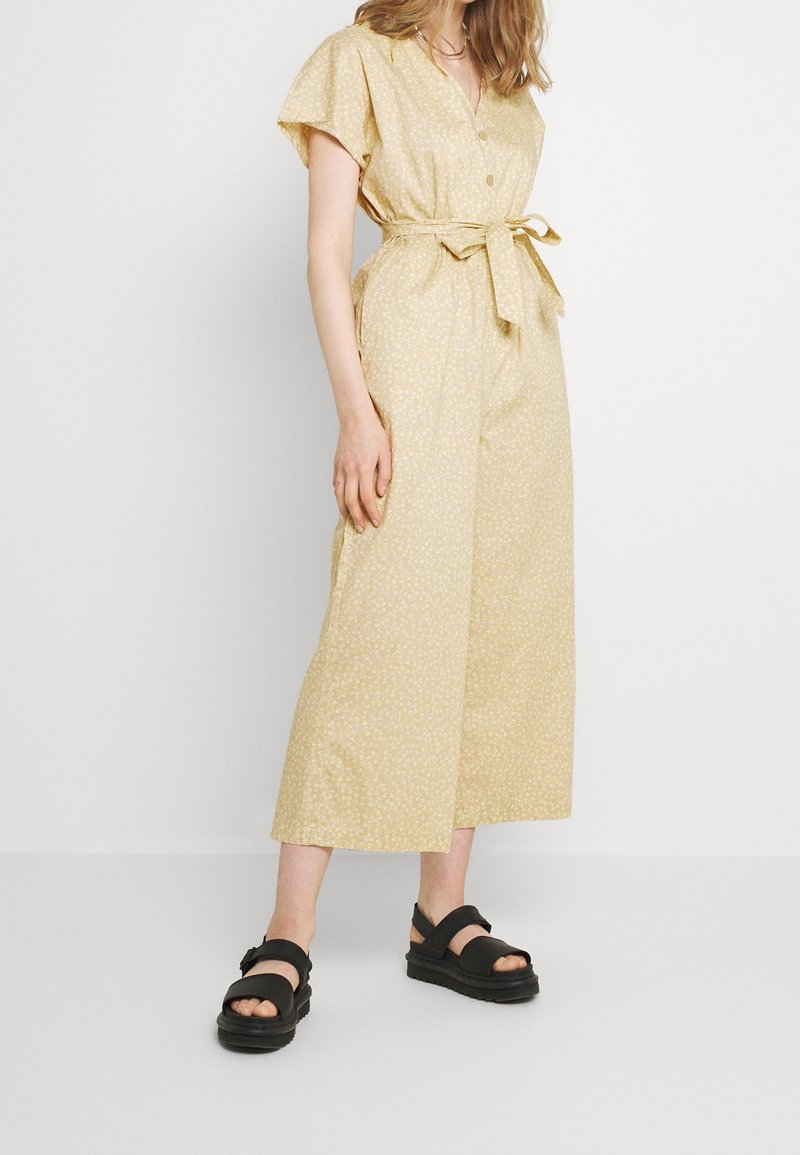 Gul blommig mönster jumpsuit med korta ärmar, en knappad v-ringning, midja med bälte och vida ben, parat med svarta sandaler.