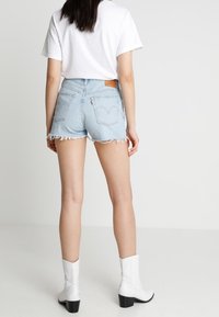 Pantalones cortos de mezclilla azul claro con dobladillo deshilachado, cintura alta y parche de cuero en la parte trasera, combinados con botas blancas hasta el tobillo. Diseño simple y casual.