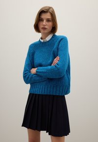 Blauwe gebreide trui met een gestructureerd patroon en geribbelde zoom, gecombineerd met een geplooide zwarte rok. De trui heeft een ronde hals en lange mouwen.