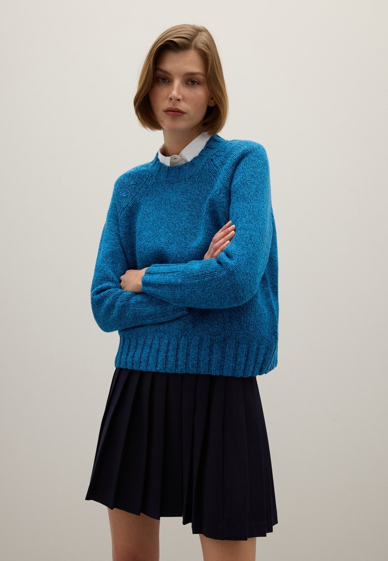 Blauwe gebreide trui met een gestructureerd patroon en geribbelde zoom, gecombineerd met een geplooide zwarte rok. De trui heeft een ronde hals en lange mouwen.