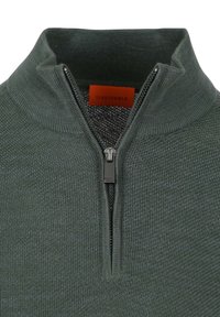 Donkergroene trui met ritssluiting, hoge kraag, gebreide textuur en een metalen rits. Voorzien van een oranje label met de tekst "SUITABLE."