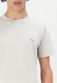 T-shirt court à manches courtes gris clair en coton. Présente un petit design brodé sur la poitrine gauche et un col rond.
