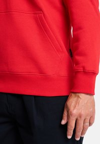 Červená mikina s klokaním vreckom, rebrovanými manžetami a lemom, vyrobená z jemného materiálu. Detailný záber na textúru a ruku ležiacu blízko.