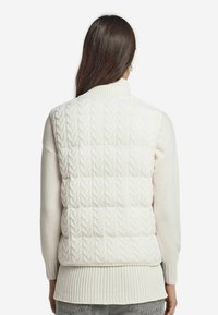 Kvinna med långt brunt hår som bär en vit kabelstickad ärmlös pufferväst över en krämfärgad tröja, sedd bakifrån.