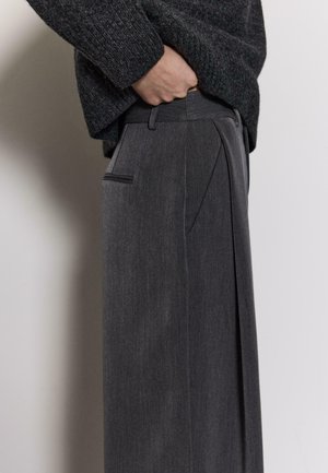 Szare plisowane spodnie o dopasowanym kroju, z kieszeniami po bokach, gładką fakturą i delikatnym połyskiem. Zestawione z ciemnym dzianinowym swetrem.