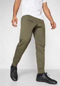 Calças de moletom verde-oliva com punhos elásticos e um logo em preto de "PARIS", combinadas com sapatos de desporto pretos com um detalhe em azul-turquesa.