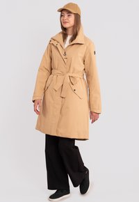 Vaaleanbeige trench-coat, jossa on vyö, vetoketju ja nepparipainikkeet. Varustettu korkealla kauluksella ja sivutaskuilla. Yhdistetty mustien housujen ja lenkkareiden kanssa.