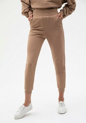 Beżowe dresowe spodnie z prążkowanym pasem, zwężanymi nogawkami, charakteryzujące się gładką, miękką fakturą oraz bocznymi kieszeniami. Połączone z białymi sneakersami.