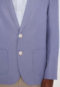 Blue blazer made of textured fabric, featuring two front pockets, three buttons, and a tailored fit. Light blue shirt underneath.