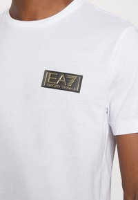 T-shirt bianca in cotone con un patch rettangolare nero e oro che riporta il testo "EA7 EMPORIO ARMANI" nella parte sinistra del petto.