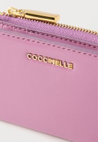 Pink leather wallet with a textured finish, featuring a gold zipper and a gold logo "Coccinelle" embossed on the surface.