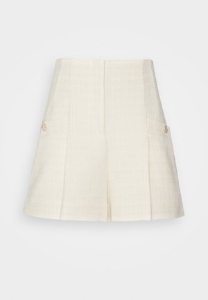 Henkilö, jolla on päällään vaaleanpunainen ruudullinen takki, valkoinen paita, kermaiset korkeavyötäröiset shortsit ja ruskeat frangeilla varustetut loaferit, seisoo puulattialla.