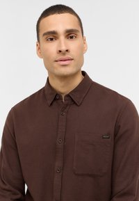 Junger Mann mit kurzen Haaren, der ein braunes, knopfbares Hemd mit einer Brusttasche trägt und frontal vor einem schlichten, hellen Hintergrund steht.